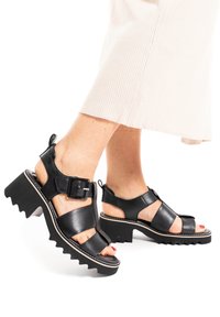 Czarne skórzane sandały z masywną, zygzakowatą podeszwą, regulowanym zapięciem i otwartym designem, z paskami na kostkach i wycięciami na palce.