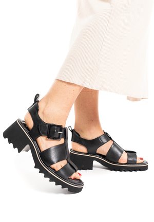 Sandali in pelle nera con suola spessa a zig zag, fibbia regolabile e design aperto, dotati di cinturini alla caviglia e ritagli sulla punta.