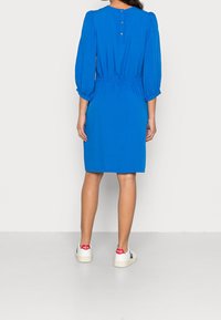 Sininen mekko, jossa on puffihihat, joustava vyötärö, takana napitusyksityiskohta ja tasaväri. Yhdistetty valkoisiin lenkkareihin, joissa on punaiset yksityiskohdat.