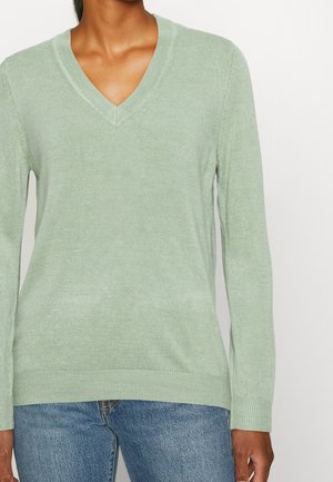 Hellgrüner Strickpullover mit V-Ausschnitt, langen Ärmeln und geripptem Saum. Der Stoff wirkt weich mit einer subtilen Textur. Getragen mit blauen Jeans.