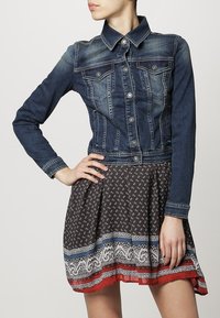 Jeansjacka med en figursydd design och två bröstfickor, kombinerad med en brun mönstrad klänning med spetskant och färgglada detaljer.