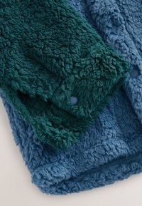 Soft textured green and blue fleece fabric close-up, showing thick, curly fibers and a hole in the green fabric layer.