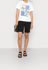 Camiseta blanca de algodón con estampados gráficos y texto verde, combinada con pantalones cortos negros y sandalias negras gruesas. Sosteniendo un bolso de mano negro.