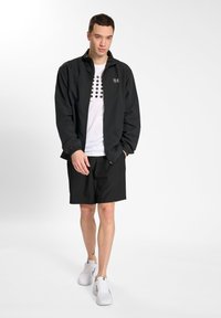 Sort vindjakke med elastiske manchetter, hvid grafisk t-shirt med sorte prikker og sorte shorts. Modellen har hvide atletiske sko på.