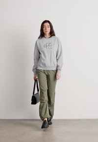 Γκρι φούτερ με λογότυπο, πράσινο cargo παντελόνι, μαύρα παπούτσια και μια μικρή μαύρη τσάντα. Το μοντέλο στέκεται μπροστά σε έναν απλό τοίχο.