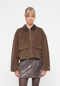 Bruine corduroyjas met een donkere kraag, ritsluiting en twee grote voorkruiserzakken. G gedragen over een paillettenrok in grijstinten en bruin.