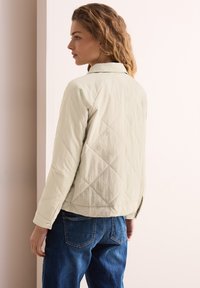 Veste matelassée beige clair à coupe décontractée, avec un col et des poignets boutonnés, associée à un jean en denim bleu.