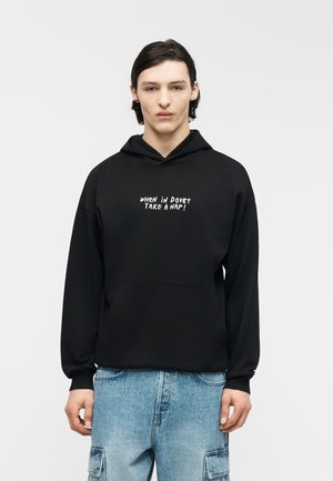 Nuori henkilö, jolla on tumma tukka, pukeutuneena mustaan huppariin, jossa lukee "WHEN IN DOUBT TAKE A NAP!", ja vaaleansinisiin cargofarkkuihin, seisoo valkoista taustaa vasten.