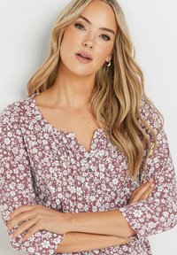 Kukkakuosiset mauve-väriset puserot, joissa on kolmannes hihoista, V-pääntie ja nappiyksityiskohta. Esittelee valkoisia kukkia teksturoidun taustan päällä.