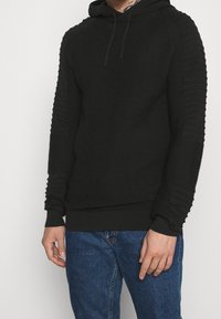 Man som bär en svart strukturerad stickad hoodie med ribbad ärmar och blå denimjeans, stående mot en enkel ljus bakgrund.