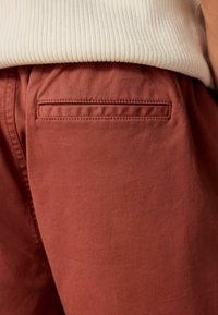 Roodbruine broek met een zachte textuur, een enkele achterzak met een horizontale opening en een relaxte fit.