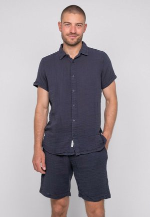 Homme aux cheveux courts portant une chemise bleu foncé à manches courtes boutonnée et un short assorti, debout avec une main dans la poche, sur un fond gris.