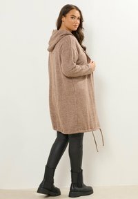 Cardigan beige con capucha hecho de tela acanalada, con un ajuste relajado y cordón en el dobladillo. Combinado con pantalones negros ajustados y botas hasta el tobillo.