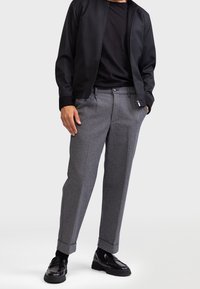 Grijze, gestructureerde broek met opgerolde pijpen, gecombineerd met een zwart jack met ritssluiting en een zwart T-shirt, afgewerkt met glanzende zwarte loafers.