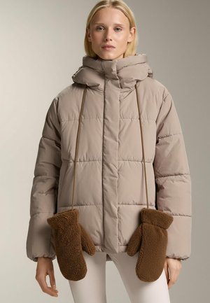 Donna che indossa una giacca imbottita beige con muffole marroni attaccate e appese a cordini, in piedi davanti a uno sfondo uniforme.