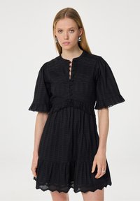 Zwarte jurk met korte pofmouwen, knoopsluiting bij de hals en een tiered ruffle zoom. Heeft een gestructureerde stof met geometrische patronen en kantaccenten.