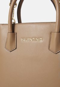 Tan spalvos odinis rankinukas su auksinėmis detalėmis, struktūrizuota forma, dvigubomis rankenomis ir iškilniu "VALENTINO" logotipu priekyje.
