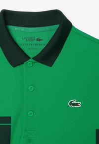 Grünes Polohemd mit schwarzem Kragen, ausgestattet mit drei passenden Knöpfen und dem Lacoste-Krokodil-Logo. Reguläre Passform, kurze Ärmel, strukturierter Stoff.