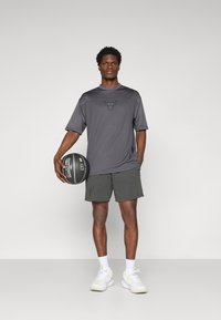Camiseta atlética de manga corta gris con un logo de los Bulls en el pecho, combinada con pantalones cortos verde oscuro y zapatillas blancas, sosteniendo un balón de baloncesto.