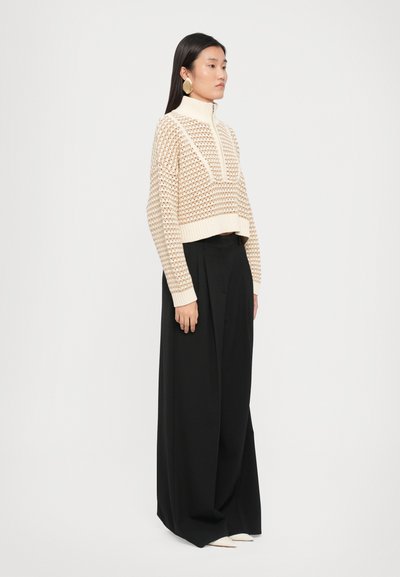 Suéter de punto color crema con cuello alto y diseño recortado, combinado con pantalones de pierna ancha negros. Tejido suave y ajuste holgado.