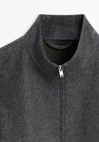 Graue Jacke zum Hochziehen mit hohem Kragen, aus strukturiertem Stoff. Verfügt über einen dezenten Glanz und einen silbernen Reißverschluss vorne.