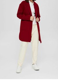 Czerwony płaszcz o luźnym kroju, z klapami z wycięciem i dwoma przednimi kieszeniami, w zestawieniu z białym t-shirtem, kremowymi spodniami i białymi trampkami.