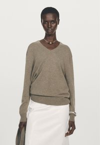 Beiger V-Ausschnitt-Pullover aus weichem Strickmaterial, mit langen Ärmeln und lockerer Passform, kombiniert mit einem weißen, fließenden Rock.
