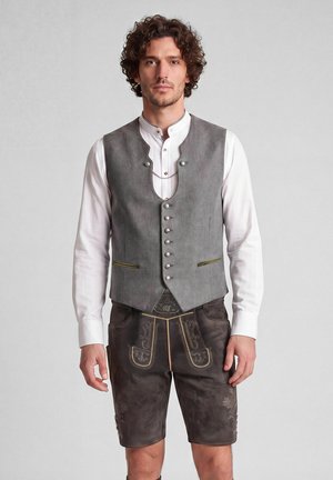 Man met krullend haar, gekleed in traditionele Beierse outfit met grijze vest, wit hemd en donkere met borduursel versierde leren korte broek, staand tegen een effen achtergrond.
