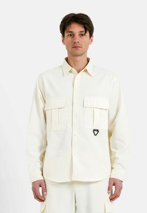 Camisa de manga larga color crema hecha de una tela texturizada, que presenta dos bolsillos en el pecho y un acento triangular negro en el bolsillo izquierdo.