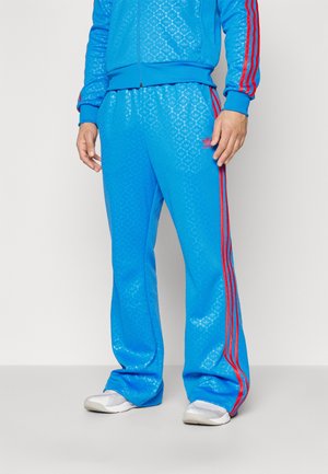 Mand iført skinnende blå Adidas træningsbukser med røde striber og logo, kombineret med hvide sneakers mod en ensfarvet baggrund.