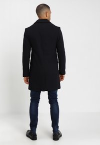 Cappotto lungo in misto lana nera con collo a scialle, vestibilità affusolata e design a lunghezza media, abbinato a jeans blu scuro e scarpe nere.