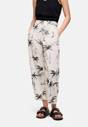 Mujer que lleva pantalones color crema con estampado de palmeras negras, top sin mangas negro y sandalias negras con suela de corcho y hebillas.