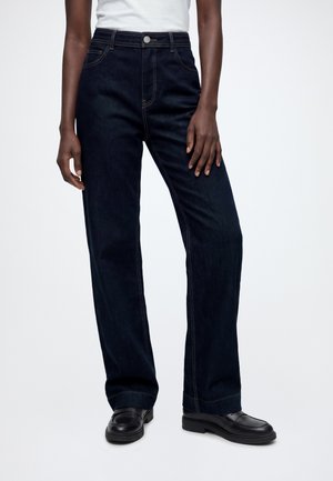 Personne portant un jean bleu foncé à jambes droites, un haut blanc et des mocassins en cuir noir, debout avec les mains détendues le long du corps.