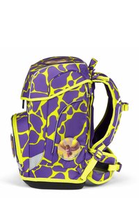 Sac à dos avec un motif de girafe violet et jaune, présentant une forme arrondie, plusieurs compartiments zippés et un badge rond sur le côté.