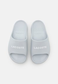 Un par de chanclas Lacoste de color azul claro con suelas texturizadas y texto del logo en blanco en las correas, vistas desde arriba sobre un fondo blanco.