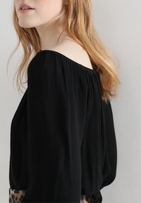 Musta olkaimeton paita, jossa on teksturoitu kangas, löysät hihat ja hulmuava malli, esitelty neutraalilla taustalla.