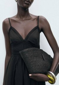 Pochette tissée noire au design incurvé, dotée d'une surface texturée et de deux fermetures éclair ; modèle portant une robe noire à bordures en dentelle.