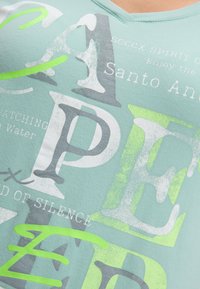 Hellgrünes V-Ausschnitt-T-Shirt mit einer Mischung aus grauen und weißen Texten sowie neon-grünen Akzenten, die verschiedene Buchstaben und Phrasen zeigen. Weicher Stoff.