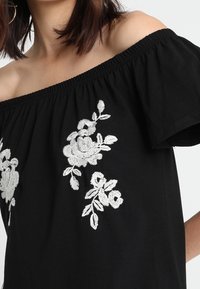 Czarny top bez ramiączek z białymi, kwiatowymi haftami. Posiada elastyczny dekolt i krótkie rękawy. Gładki bawełniany materiał.