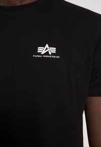 Czarna koszulka z małym białym logo Alpha Industries i napisem umiejscowionym na środku klatki piersiowej, noszona przez osobę o ciemnej karnacji.