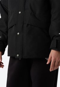 Černá nepromokavá bunda s patentními knoflíky, bočními kapsami a texturovanou látkou. Rukáv zdobí logo; doplněno černými kalhotami.