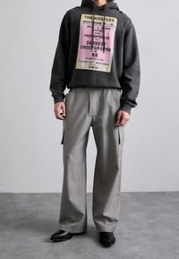 Felpa con cappuccio di colore grigio scuro con stampa grafica, abbinata a pantaloni grigi larghi con tasche laterali e una texture liscia. Scarpe nere.