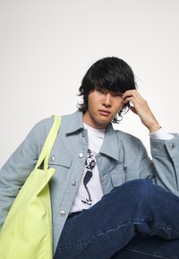Chaqueta de mezclilla azul clara con botones plateados, camiseta gráfica blanca, jeans oscuros y una tote bag amarilla brillante. El cabello es oscuro y ondulado.
