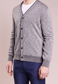 Man i en ljusgrå knappad cardigan över en vit T-shirt och mörkblå byxor, stående mot en enkel bakgrund.