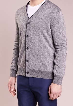 Uomo che indossa un cardigan grigio chiaro con bottoni sopra una t-shirt bianca e pantaloni blu scuri, in piedi contro uno sfondo semplice.