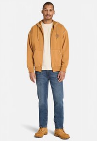 Beżowa bluza z kapturem z przednimi kieszeniami, noszona na kremowej koszulce, w parze z niebieskimi dżinsami i żółtymi butami roboczymi.