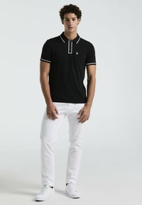 Schwarzes Poloshirt mit weißen Akzenten, kurzen Ärmeln und einer kleinen Brusttasche. Kombiniert mit schmal geschnittenen weißen Hosen und weißen Sneakers.
