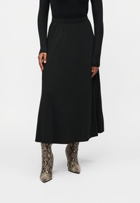 Μαύρη μίντι φούστα με λεία υφή, με εφαρμοστό ζωνάκι στη μέση και φουσκωτή σιλουέτα. Συνδυάζεται με ψηλές μπότες μέχρι το γόνατο με σχέδιο φίδι.