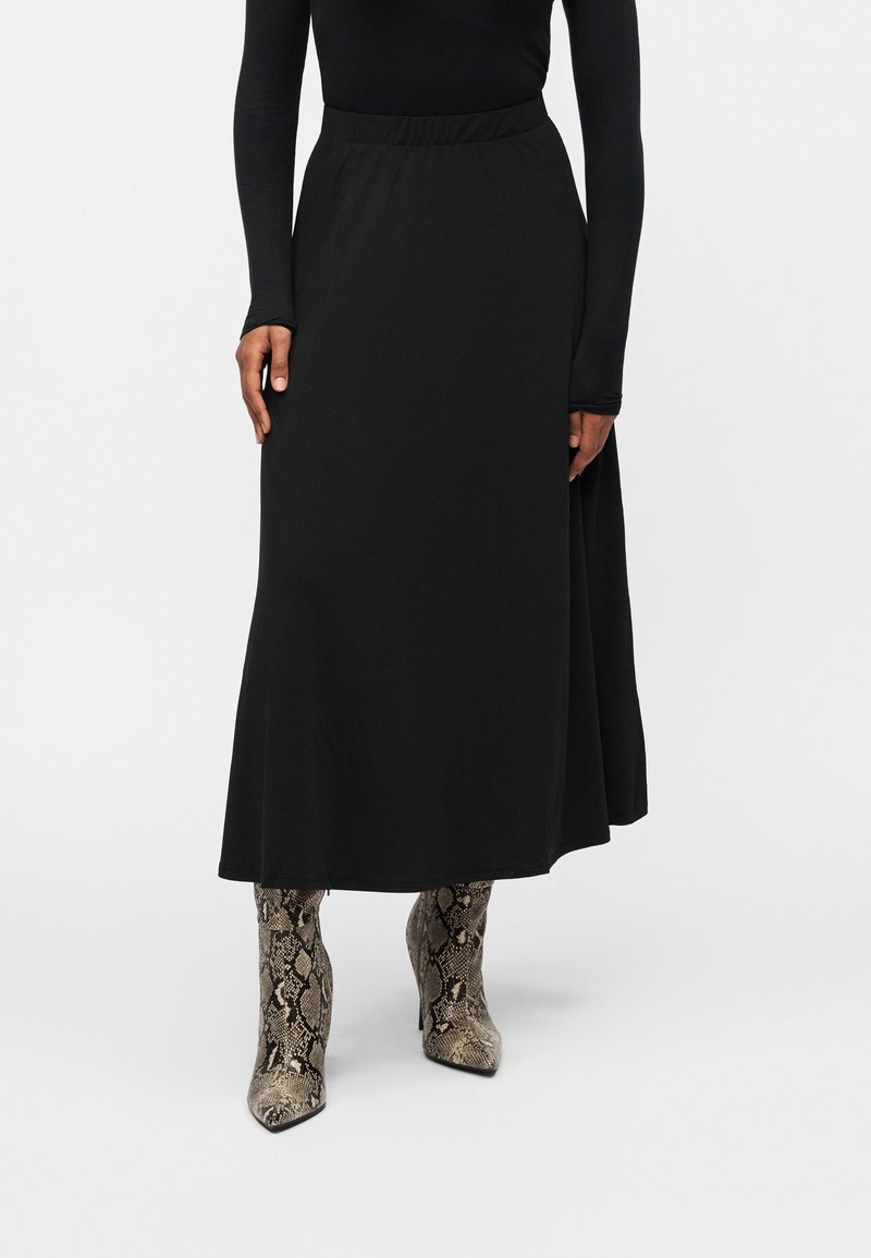 Μαύρη μίντι φούστα με λεία υφή, με εφαρμοστό ζωνάκι στη μέση και φουσκωτή σιλουέτα. Συνδυάζεται με ψηλές μπότες μέχρι το γόνατο με σχέδιο φίδι.