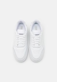 Paire de baskets blanches Puma avec lacets, logo sur la languette et zone perforée au niveau des orteils, vue de dessus sur un fond uni.